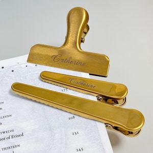K&ouml;nnte beinhalten: Drei goldfarbene Metall-Ordnerklammern, jeweils mit dem Namen "Catherine" graviert. Die Klammern sind in verschiedenen Gr&ouml;&szlig;en erh&auml;ltlich, mit einer gro&szlig;en Klammer oben und zwei kleineren darunter. Die Klammern liegen auf einer Seite mit Text.
