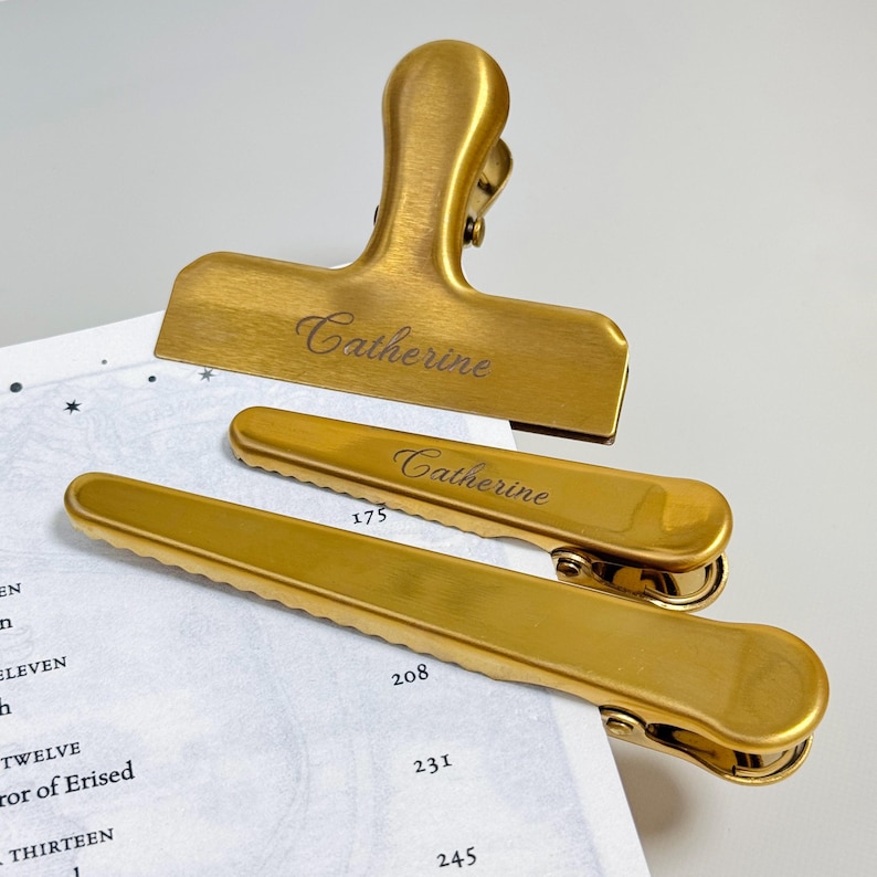 K&ouml;nnte beinhalten: Drei goldfarbene Metall-Ordnerklammern, jeweils mit dem Namen "Catherine" graviert. Die Klammern sind in verschiedenen Gr&ouml;&szlig;en erh&auml;ltlich, mit einer gro&szlig;en Klammer oben und zwei kleineren darunter. Die Klammern liegen auf einer Seite mit Text.