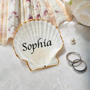 Puede incluir: Un plato decorativo en forma de concha de vieira blanca con borde dorado y el nombre Sophia impreso en letras cursivas negras. La concha se acompaña de dos anillos de metal plateado y una pequeña perla blanca sobre una tela de encaje.