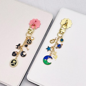 Puede incluir: Dos llaveros de metal dorado con múltiples dijes. El llavero izquierdo tiene una base rosa en forma de flor, una letra H, una llave, un corazón, una luna creciente y un colgante ovalado de rosa. El derecho tiene una base de flor dorada, una estrella, un planeta, una flor y una luna creciente azul y verde.