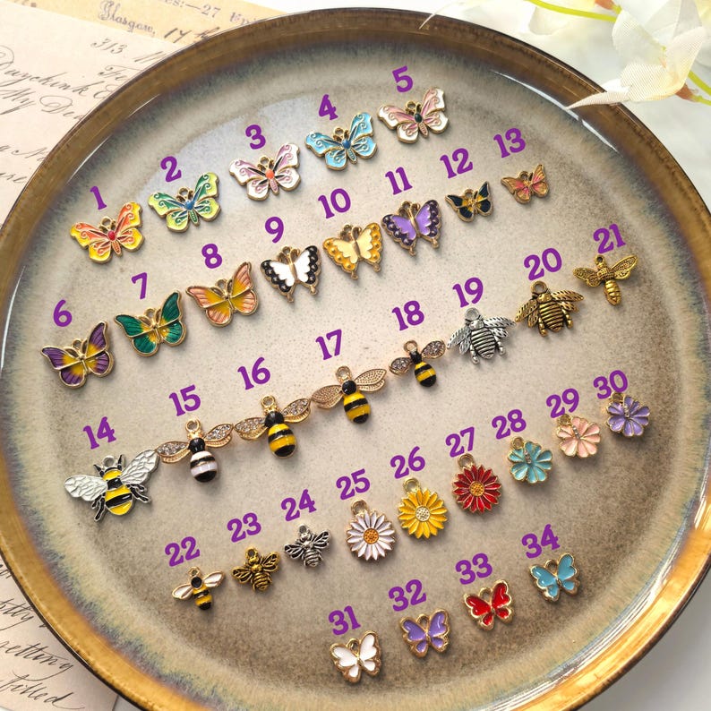 May include: A collection of small, colorful charms in various designs, including butterflies, bees, and flowers. The charms are arranged on a round, brown-toned plate. Each charm has a unique color scheme and intricate details.