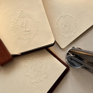 May include: Three cream-coloured paper cards featuring embossed monograms and names. One card shows the letter L and Lucas, another shows RJ and RYAN JAMES, and the third shows AM and AIDEN MILLER. A metal hand embosser tool sits to the right.