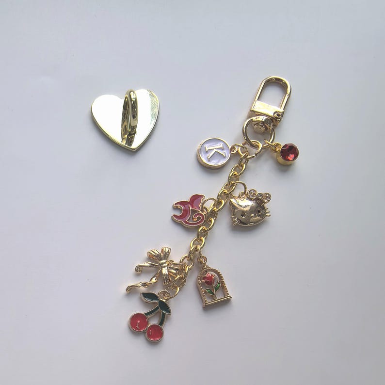 Puede incluir: Un llavero dorado con m&uacute;ltiples colgantes, incluyendo una cabeza de Hello Kitty, una gema roja, una rosa en una jaula, cerezas y un lazo. Tambi&eacute;n hay un gancho dorado en forma de coraz&oacute;n separado.
