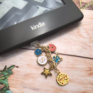 Puede incluir: Una pulsera de dijes dorada con varios dijes de esmalte, que incluyen una flor azul, un corazón rosa, un círculo blanco con una "S", un planeta, una estrella y un dije "Lucky". El dije está unido a un lector Kindle.