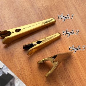 May include: Three gold-colored metal clips in different styles, arranged on a wooden surface. The clips are labeled "Style 1", "Style 2", and "Style 3". The clips have a spring-loaded design and a serrated edge.