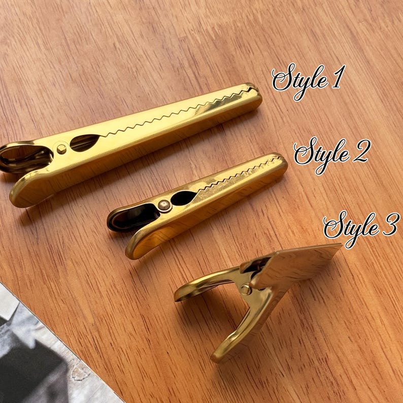 May include: Three gold-colored metal clips in different styles, arranged on a wooden surface. The clips are labeled "Style 1", "Style 2", and "Style 3". The clips have a spring-loaded design and a serrated edge.