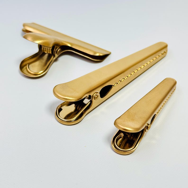 K&ouml;nnte beinhalten: Drei goldfarbene Metall-Ordnerklammern in verschiedenen Gr&ouml;&szlig;en sind auf einer wei&szlig;en Oberfl&auml;che angeordnet. Die Klammern haben eine matte Oberfl&auml;che und ein klassisches Design mit Federmechanismus zum Sichern von Papieren oder anderen Gegenst&auml;nden.
