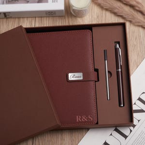 Op de afbeelding: Een bordeauxrood leren notitieboek en pennenset in een geschenkdoos. Het notitieboek heeft een zilveren naamplaatje met "Ross" en initialen "R&S". Een zilveren penvulling en een bordeauxrode pen zijn inbegrepen.