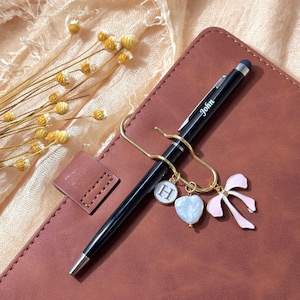 May include: A black pen, inscribed with "John", held by a gold pen clip. The clip features charms: a white "H", a heart-shaped charm, and a pink bow. The pen rests on a brown leather notebook, with a few dried flowers.