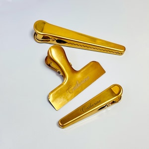 K&ouml;nnte beinhalten: Drei goldfarbene Metall-Beutelclips in verschiedenen Gr&ouml;&szlig;en. Der gr&ouml;&szlig;te Clip ist rechteckig, der mittlere Clip ist T-f&ouml;rmig und der kleinste Clip ist rechteckig. Der Name "Catherine" ist auf dem mittleren und kleinsten Clip eingraviert.