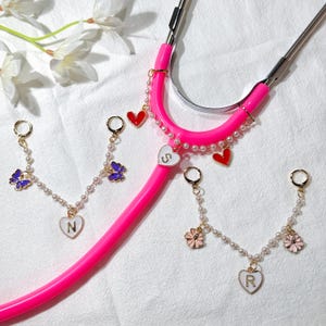 Peut inclure: Un stéthoscope rose vif orné de chaînes de perles et de breloques. Les breloques comprennent des cœurs rouges, des pendentifs en forme de cœur blancs avec des lettres et des papillons et fleurs dorés. Les parties métalliques du stéthoscope sont argentées.