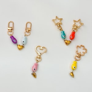 Puede incluir: Una colección de llaveros coloridos con herrajes dorados. Cada llavero presenta un diseño único, que incluye un corazón, una estrella y un trébol. Los dijes están adornados con cuentas vibrantes en tonos morado, azul, rojo, amarillo y rosa, con detalles dorados.