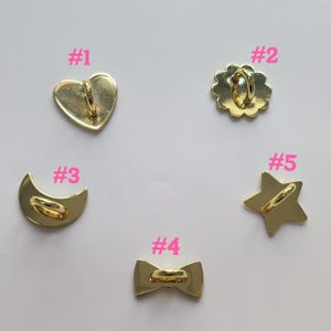 Puede incluir: Cinco dijes de metal dorado en varias formas: un coraz&oacute;n, una flor, una media luna, una pajarita y una estrella. Cada dije tiene un lazo en la parte posterior para sujetarlo. Los dijes se muestran sobre una superficie blanca.
