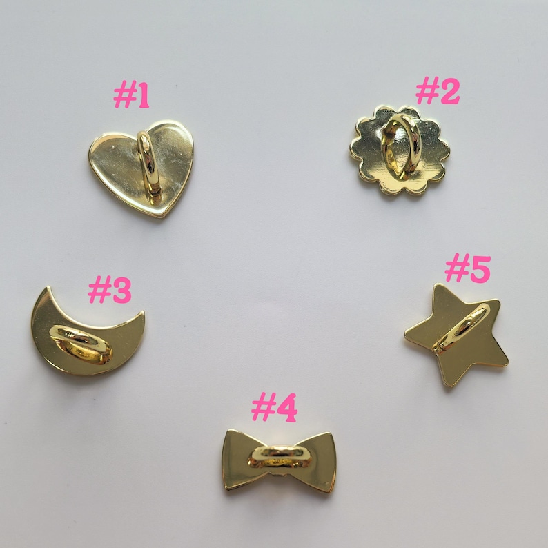 Puede incluir: Cinco dijes de metal dorado en varias formas: un coraz&oacute;n, una flor, una media luna, una pajarita y una estrella. Cada dije tiene un lazo en la parte posterior para sujetarlo. Los dijes se muestran sobre una superficie blanca.