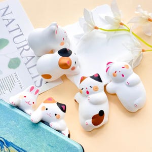 Può includere: Una collezione di statuine in ceramica bianca a forma di gatti e un coniglio. Le statuine hanno dettagli neri, marroni e rosa. Alcune sono sdraiate, altre in piedi. Le statuine sono su una superficie bianca con una composizione floreale.