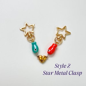 Op de afbeelding: Een goudkleurige metalen sluiting met twee stervormige uiteinden. De sluiting heeft een turquoise en rode visvormige bedel, verbonden door een gouden hart. De tekst "Style 2 Star Metal Clasp" staat eronder.