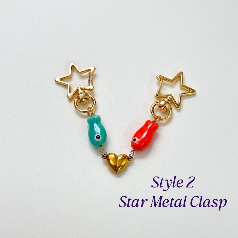 Op de afbeelding: Een goudkleurige metalen sluiting met twee stervormige uiteinden. De sluiting heeft een turquoise en rode visvormige bedel, verbonden door een gouden hart. De tekst "Style 2 Star Metal Clasp" staat eronder.