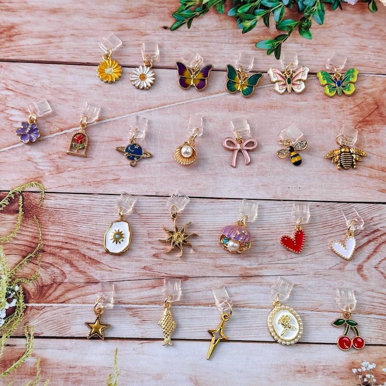 May include: A collection of decorative earring clips with various designs, including flowers, butterflies, bees, hearts, and stars. The clips are gold-toned with colorful enamel details and clear plastic attachments.