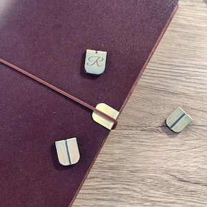 Puede incluir: Un diario de cuero burdeos con cierre elástico marrón y tres marcadores de página metálicos. Dos marcadores son rectangulares con una raya vertical, y uno tiene la letra "R". El diario está sobre una superficie de madera.