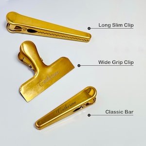 K&ouml;nnte beinhalten: Drei goldfarbene Metallclips: ein langer, schlanker Clip, ein Clip mit breitem Griff und ein klassischer Bar-Clip. Der Clip mit breitem Griff und der klassische Bar-Clip tragen den eingravierten Namen "Catherine". Das Bild ist auf wei&szlig;em Hintergrund.