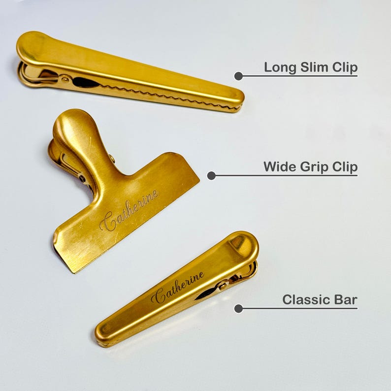 K&ouml;nnte beinhalten: Drei goldfarbene Metallclips: ein langer, schlanker Clip, ein Clip mit breitem Griff und ein klassischer Bar-Clip. Der Clip mit breitem Griff und der klassische Bar-Clip tragen den eingravierten Namen "Catherine". Das Bild ist auf wei&szlig;em Hintergrund.