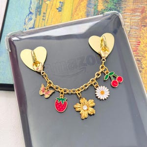 May include: Gold-coloured heart-shaped phone grip with a charm chain featuring a butterfly, strawberry, flower, daisy, and cherries. The grip is attached to a clear phone case with the Amazon logo.