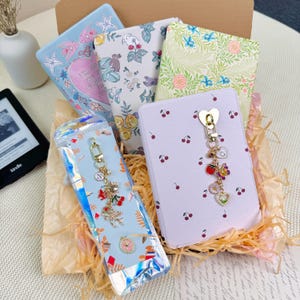 Könnte beinhalten: Eine Geschenkbox mit dekorativen Notizbüchern und einem Charm-Schlüsselanhänger. Die Notizbücher zeigen florale, herzförmige und maritime Designs. Der Schlüsselanhänger hat verschiedene Charms, darunter eine Erdbeere, ein Schmetterling und eine Blume.