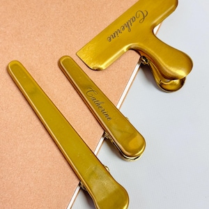 K&ouml;nnte beinhalten: Drei goldfarbene Metallclips, darunter zwei lange, schlanke Clips und ein gr&ouml;&szlig;erer Clip mit Griff. Der Name "Catherine" ist auf zwei der Clips eingraviert. Die Clips sind auf einer rosa und wei&szlig;en Oberfl&auml;che angeordnet.