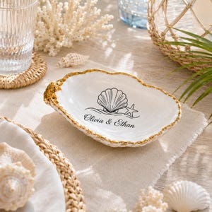 Peut inclure: Un vide-poche en forme de coquille d'huître blanche avec un bord festonné peint en doré. L'intérieur présente une illustration au trait noir d'un coquillage et d'une étoile de mer, avec le texte "Olivia & Ethan" imprimé sous le motif.