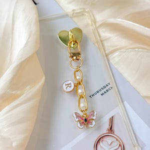 Könnte beinhalten: Goldfarbener Handy-Anhänger mit herzförmigem Verschluss und einer Kette mit Perlen und einem Schmetterlingsanhänger. Der Schmetterling ist weiß, rosa und grün. Ein Anhänger mit dem Buchstaben "R" ist ebenfalls enthalten. Der Anhänger ist an einer transparenten Handyhülle befestigt.