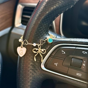 May include: A gold-colored car charm with a white heart pendant featuring the letter "G", a bow, and a blue gem. The charm is attached to a black steering wheel.