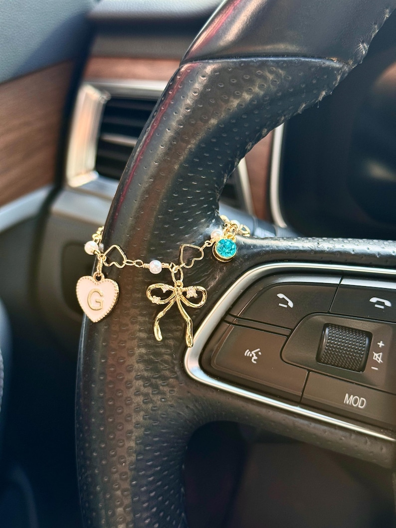 May include: A gold-colored car charm with a white heart pendant featuring the letter "G", a bow, and a blue gem. The charm is attached to a black steering wheel.