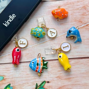 Op de afbeelding: Een verzameling kleurrijke visvormige bedels, elk met een uniek ontwerp en kleurenschema, waaronder rood, blauw, oranje en geel. Sommige bedels zijn gecombineerd met goudkleurige initiaalhangers. Het woord "kindle" is zichtbaar op een zwart apparaat.