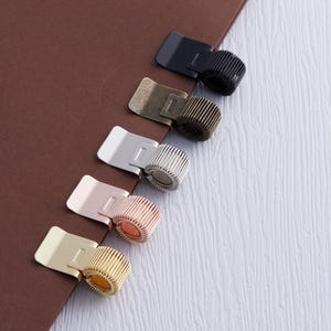 May include: A collection of pen holders in various metallic finishes, including black, brass, silver, rose gold, and gold. Each holder features a rectangular clip and a ribbed ring to secure a pen or pencil. The holders are arranged on a brown and white surface.