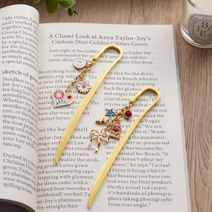 May include: Two gold-colored metal bookmarks with decorative charms. One bookmark features a heart, daisy, and circle charm. The other has a bow, key, star, and flower charm. Both are on a book with text.