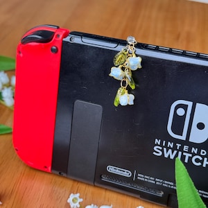 Pu&ograve; includere: Un delicato ciondolo floreale con pendenti a forma di fiori blu pallido e foglie verdi &egrave; appeso a una console Nintendo Switch. Il ciondolo &egrave; dorato e la console &egrave; nera e rossa. Il logo Nintendo Switch &egrave; visibile.