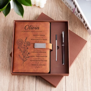 Peut inclure: Un journal en cuir marron avec le nom "Olivia" et un texte inspirant, présenté dans un coffret cadeau avec un stylo assorti et un stylo argenté. Le journal présente un motif floral et un fermoir en métal. La boîte est marron.