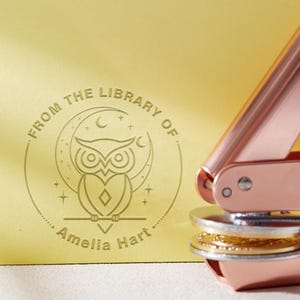 Op de afbeelding: Een roségouden en zilveren zegelstempel naast een gouden kaart met een reliëf uilontwerp. De kaart leest "From the Library of Amelia Hart" in een cirkelvormig patroon rond de uilillustratie.