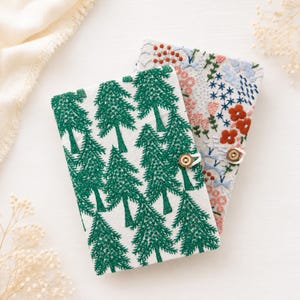 May include: Two rectangular fabric-covered notebooks with button closures. The top notebook features a white background with a repeating green pine tree pattern. The bottom notebook has a white background with floral embroidery in red, pink, and blue.