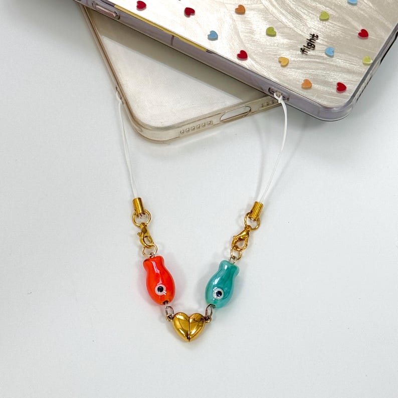Op de afbeelding: Een witte telefoonlanyard met goudkleurige sluitingen en een bedel met een gouden hart en twee visvormige kralen, &eacute;&eacute;n rood en &eacute;&eacute;n blauwgroen. De telefoonhoes is transparant met een hartjespatroon.
