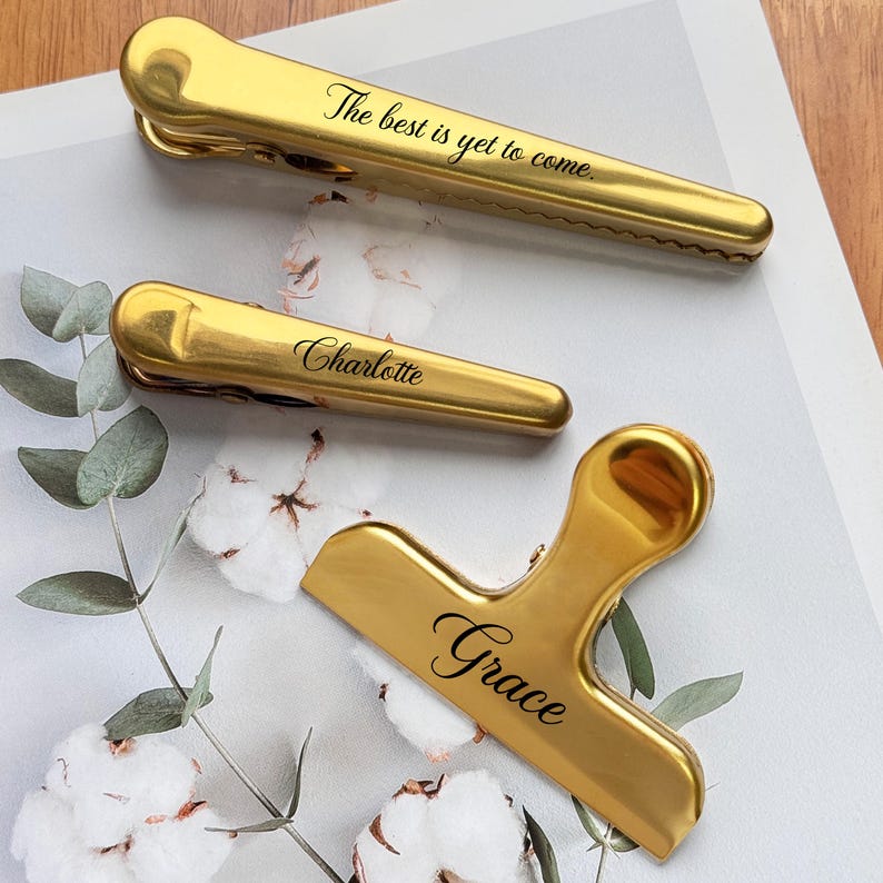 May include: Three gold-colored bag clips with black text. One clip reads "The best is yet to come." Another reads "Charlotte," and the third reads "Grace." The clips are on a white and light green floral background.
