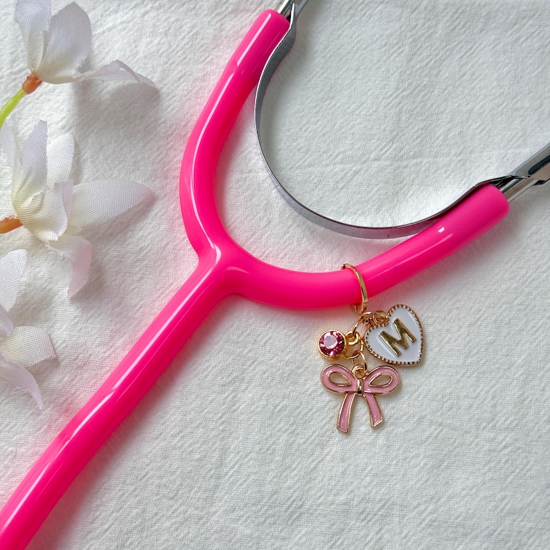 Pink stethoscope with custom charms including a pink bow, heart initial charm, and rhinestone pendant — personalized medical accessory gift for nurses.
