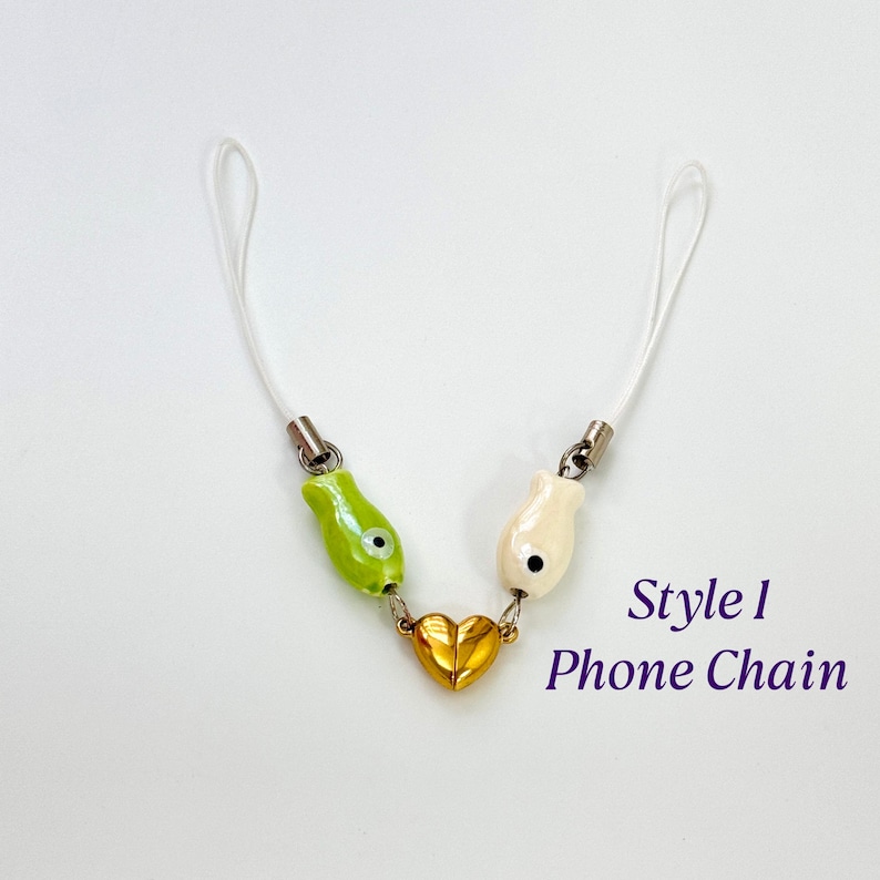 Op de afbeelding: Een telefoonketting met een wit koord, met een groene en cr&egrave;mekleurige bedel, verbonden door een gouden hart. De tekst "Style 1 Phone Chain" is zichtbaar in de afbeelding.