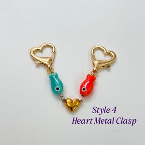 Op de afbeelding: Een goudkleurige hartvormige sluiting met twee hartvormige sluitingen en twee visvormige bedels, &eacute;&eacute;n turquoise en &eacute;&eacute;n rood. Een gouden hartbedel verbindt de visbedels. De tekst "Style 4 Heart Metal Clasp" staat eronder.