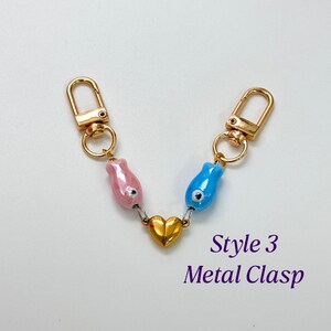 Op de afbeelding: Een goudkleurige metalen sluiting met twee karabijnsluitingen verbonden door een ketting. De ketting heeft een roze en een blauwe kraal en een gouden hartbedel. De tekst "Style 3 Metal Clasp" is zichtbaar.