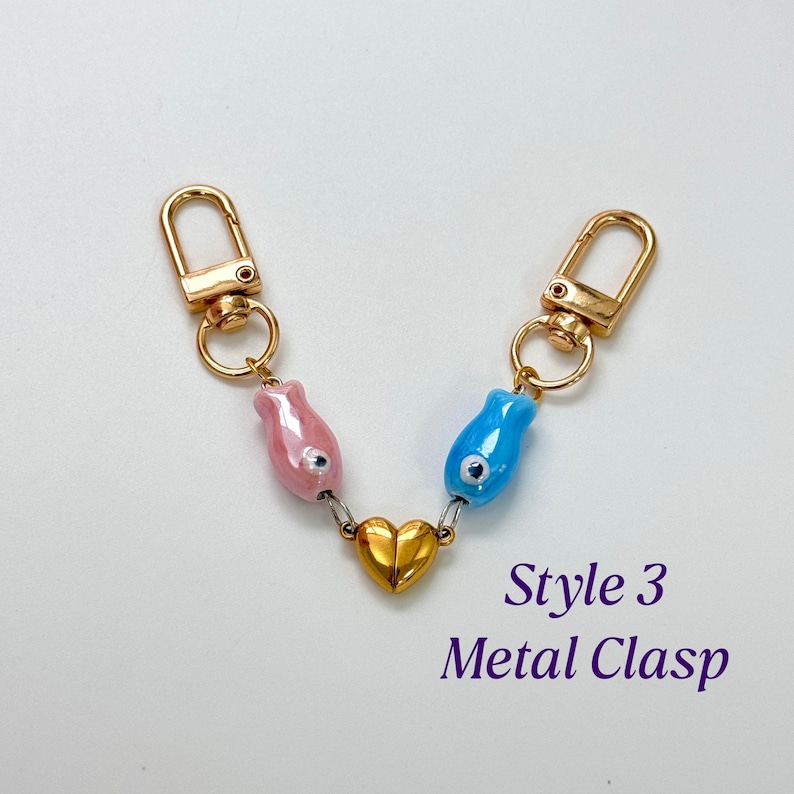 Op de afbeelding: Een goudkleurige metalen sluiting met twee karabijnsluitingen verbonden door een ketting. De ketting heeft een roze en een blauwe kraal en een gouden hartbedel. De tekst "Style 3 Metal Clasp" is zichtbaar.