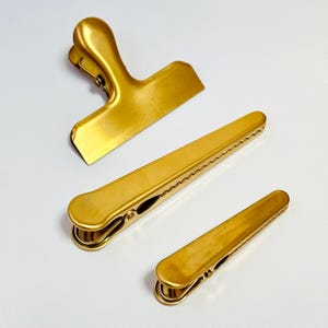 K&ouml;nnte beinhalten: Drei goldfarbene Metall-Beutelclips in verschiedenen Gr&ouml;&szlig;en. Der gr&ouml;&szlig;te Clip hat einen Griff, w&auml;hrend die anderen beiden lang und schmal sind. Die Clips sind auf einer wei&szlig;en Oberfl&auml;che angeordnet.