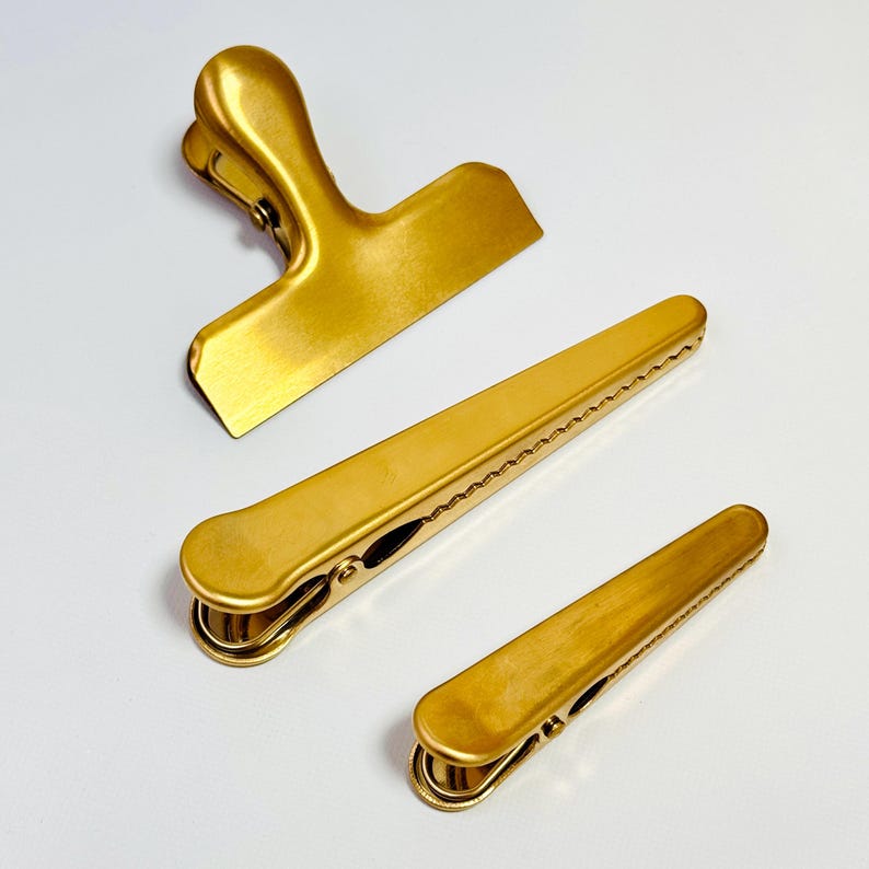 K&ouml;nnte beinhalten: Drei goldfarbene Metall-Beutelclips in verschiedenen Gr&ouml;&szlig;en. Der gr&ouml;&szlig;te Clip hat einen Griff, w&auml;hrend die anderen beiden lang und schmal sind. Die Clips sind auf einer wei&szlig;en Oberfl&auml;che angeordnet.
