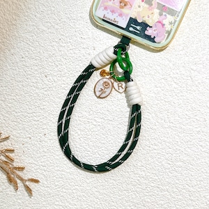 Op de afbeelding: Groene en witte telefoonbedel met een goudkleurige rozenhanger en de letter "R". De bedel heeft een groen-wit gestreept koord, witte accenten en een groene ring. De telefoonhoes heeft een konijnenontwerp.