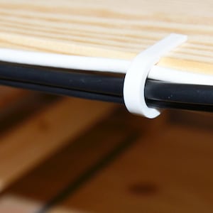 May include: White plastic cable clip holding two black cables against a light wood surface.